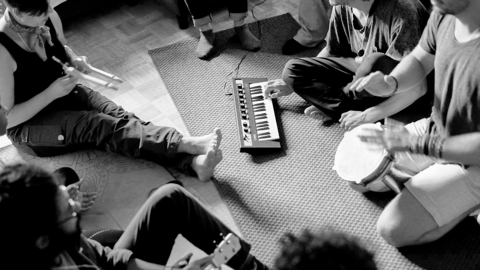 a group seated in a circle on the floor, playing a small keyboard, hand drums and other instruments together